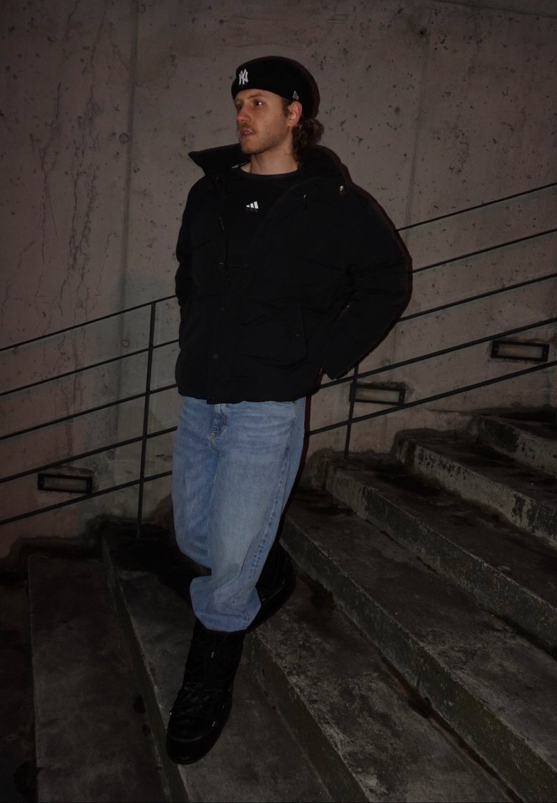 Jeune homme en veste noire, jean bleu et casquette noire appuyé sur une rambarde à côté d'escaliers en béton la nuit.