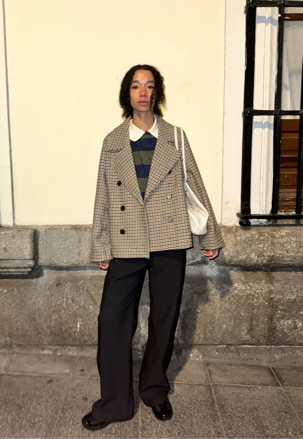 Blazer surdimensionné à carreaux dans des tons marron et crème, associé à un polo rayé bleu et vert. Pantalons larges noirs et un sac blanc.