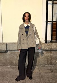 Blazer surdimensionné à carreaux dans des tons marron et crème, associé à un polo rayé bleu et vert. Pantalons larges noirs et un sac blanc.