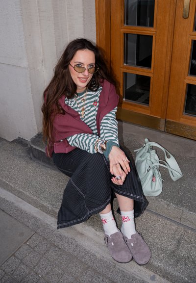 Joven sentada en escalones de piedra junto a una puerta de madera, lleva una camisa de rayas, falda oscura, gafas de sol, varias pulseras y zapatos claros con calcetines.
