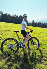 Ezüst vázú hegyikerékpár, fekete gumikkal és sportos kialakítással. A kerékpáros fehér pólót és fehér sisakot visel, amelynek tükröződő napellenzője van.