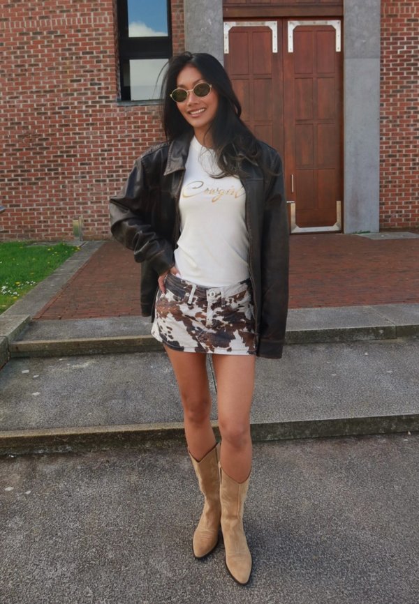 Mlada ženska z okroglimi sončnimi očali, črno usnjeno jakno, belo majico z grafiko, rjavo-belo vzorčasto mini krilo in svetlo rjavimi kavbojskimi škornji na prostem.