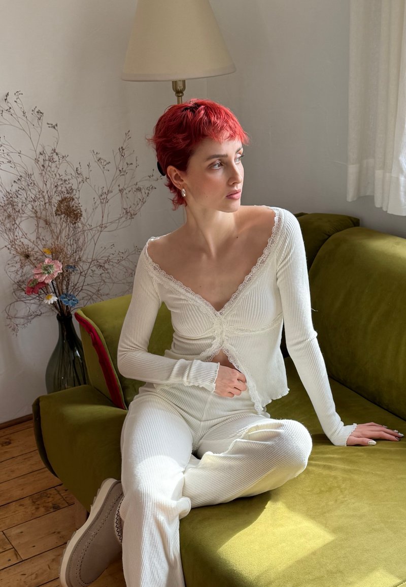 Person mit kurzen roten Haaren, der ein weißes, geripptes Oberteil und eine Hose trägt und auf einem olivgrünen Sofa in einem sanft beleuchteten Raum mit getrockneten Blumen und Lampe sitzt.