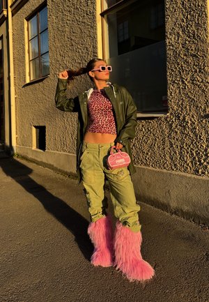 Veste imperméable verte sur un haut à motifs léopard roses, pantalon cargo et bottes roses en fausse fourrure. Petit sac à main de marque rose. Fond urbain ensoleillé.