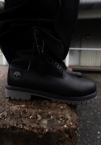 Botte en cuir noir avec semelle grise épaisse, logo en métal et étiquette chaîne posée sur une surface en pierre rugueuse en plein air.