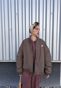 Καφέ φουσκωτό μπουφάν με ραβδωτή υφή, σε συνδυασμό με φούστα από καμπαρτίνα. Ένα κασκόλ με σχέδια κοσμεί το κεφάλι, με χρυσά σκουλαρίκια ορατά.