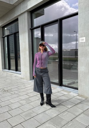 Mujer que lleva un jersey con cremallera morado, pantalones grises de pierna ancha, botas negras de tacón, gafas de sol y gorra, de pie frente a un edificio moderno con grandes ventanas.