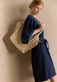 Beige strukturierte Tote-Tasche mit zwei weißen Trägern, rechteckig geformt und mit einem kleinen Vorderetikett. Das Model trägt ein lockeres, navy-blaues Kleid.