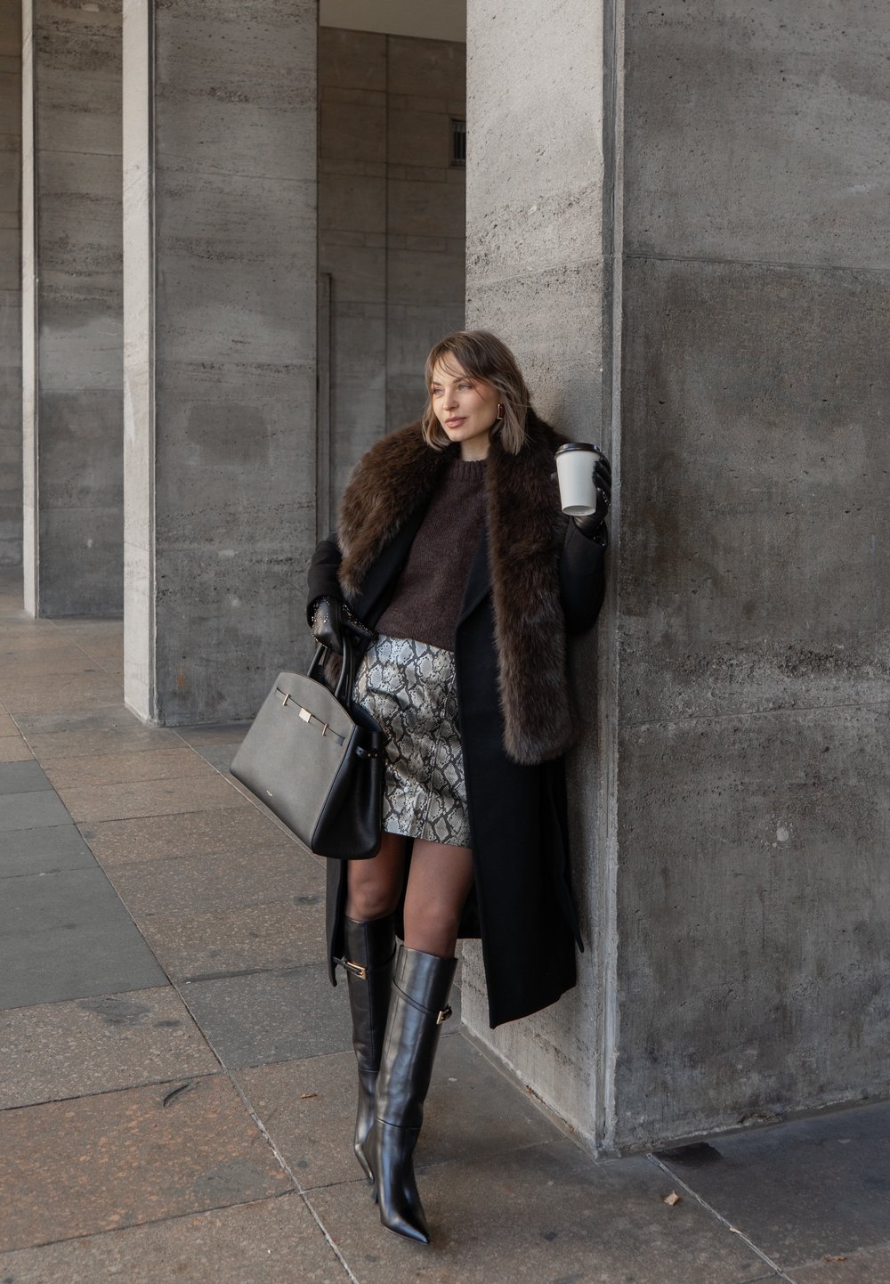 Žena v čiernom kabáte s kožušinovým golierom, metalickou sukňou s hadím vzorom, čiernymi čižmami po kolená, držiaca pohár kávy, pred betónovou stenou.