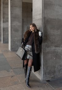 Žena v čiernom kabáte s kožušinovým golierom, metalickou sukňou s hadím vzorom, čiernymi čižmami po kolená, držiaca pohár kávy, pred betónovou stenou.