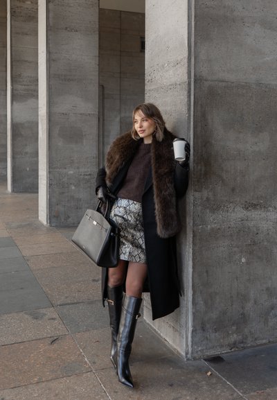 Mujer con abrigo negro con cuello de piel sintética, falda metálica con estampado de serpiente, botas negras hasta la rodilla, sosteniendo una taza de café, frente a una pared de hormigón.