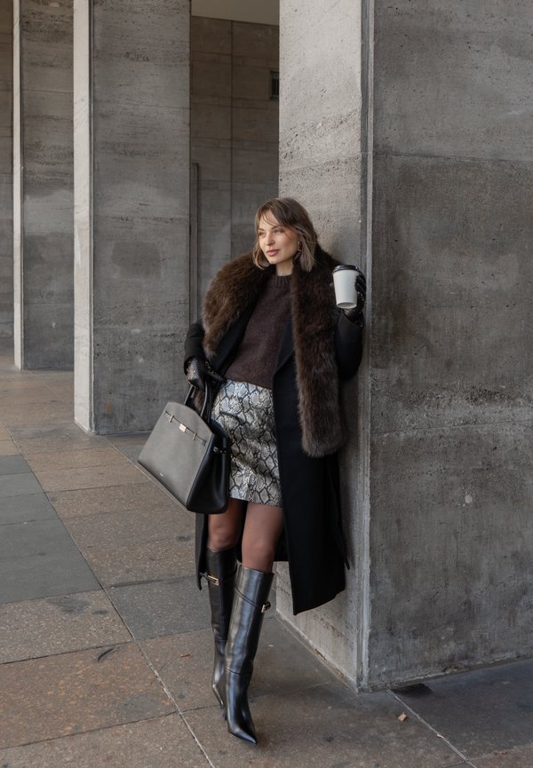 Femme en manteau noir avec un col en fausse fourrure, jupe métallisée à motif serpent, bottes noires jusqu'aux genoux, tenant une tasse de café, contre un mur en béton.