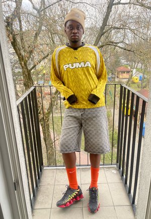 Camisa de manga larga amarilla de PUMA con puños negros, pantalones cortos grises con estampado, calcetines naranjas y zapatillas negras y rojas de Nike en un balcón.
