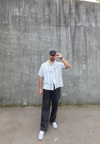 Camisa de cuadros de manga corta en azul claro y blanco, combinada con pantalones sueltos negros y zapatillas grises. Sujeto con una gorra negra.