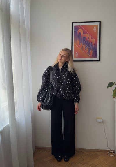 Mujer con cabello largo y rubio que lleva una blusa negra con lunares y pantalones negros anchos, de pie cerca de una ventana con un póster enmarcado y colorido en la pared.
