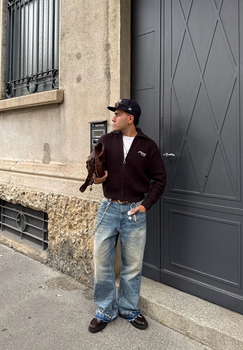 Pull à zipper en laine noire, jeans délavés bleu ample et chaussures brunes. Tient un sac marron. Mur gris et porte sombre en arrière-plan.