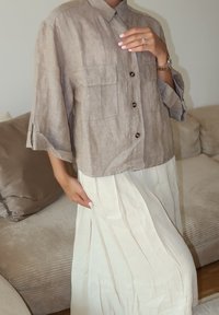 Pamučni košulja s dugim rukavima u svijetlosmeđoj boji s dva džepa na prsima, u kombinaciji s dugom, plisiranom bijelom suknjom, koja prikazuje opušteni kroj i kontrast tekstura.
