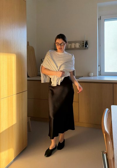 Mujer con gafas, blusa blanca, suéter gris sobre los hombros y falda negra caminando en una cocina iluminada por el sol con armarios de madera.