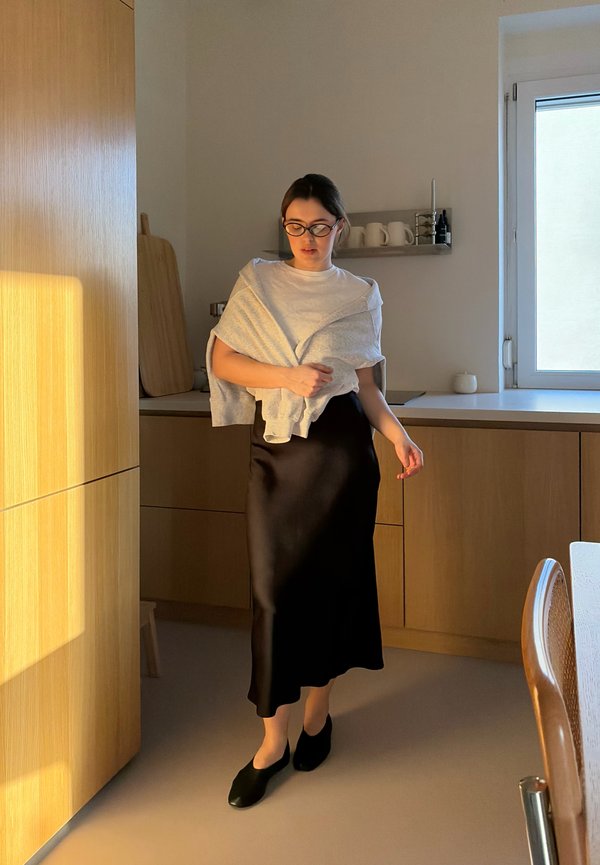 Vrouw met een bril, wit shirt, een grijze trui over haar schouders gedrapeerd, en een zwarte rok, lopend in een door zonlicht verlichte keuken met houten kasten.