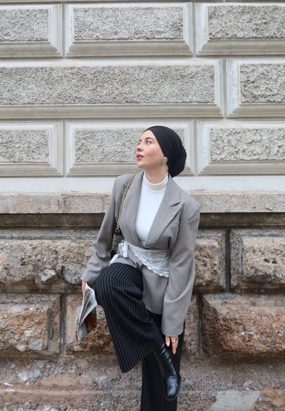 Mujer con pañuelo negro en la cabeza y blazer gris con cinturón de encaje, pantalones de rayas, sosteniendo un periódico, ajustándose una bota negra junto a una pared de piedra.