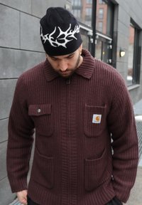 Homme portant un pull zippé côtelé bordeaux avec poches et un bonnet noir orné d'un motif de bois de cerf blanc, regardant vers le bas à l'extérieur près d'un bâtiment gris.