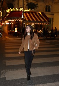 Brun mockajacka bomber, krämfärgad tröja, mörka byxor, svarta stövlar med klack. Gatubild med café i bakgrunden, randig markis, varma ljus.