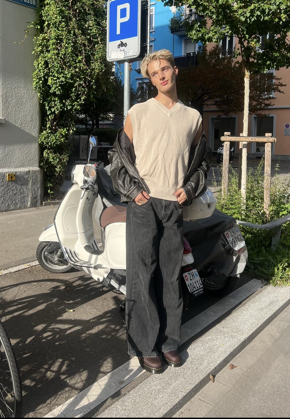 Jeune personne portant un pull sans manches et une veste en cuir, debout à côté d'un scooter blanc garé sous un panneau de stationnement dans un environnement urbain ensoleillé.