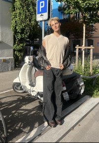 Jeune personne portant un pull sans manches et une veste en cuir, debout à côté d'un scooter blanc garé sous un panneau de stationnement dans un environnement urbain ensoleillé.
