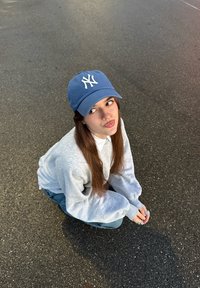 Casquette de baseball bleue avec logo blanc, sweat-shirt gris et jean bleu. Le sujet est accroupi sur l'asphalte, les mains jointes au niveau des genoux.