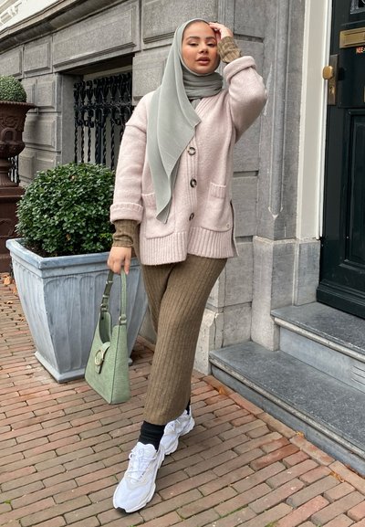 Cárdigan de punto rosa claro con grandes botones, pantalones de canalé olivo, zapatillas deportivas blancas gruesas y un bolso verde. Un hiyab gris cae con estilo.