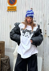 Juodas pūkuotas švarkas, baltas marškinėliai su užrašu "BLONDIE" ir laisvi juodi kelnes. Mėlynas kepurė su tekstu. Fone matosi ženklas ir mediena.
