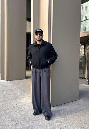 Schwarze Zip-Jacke, graue weite Hose, schwarze Sneaker und Baseballcap mit Text. Steht gegen eine beige Säule und Glastüren.