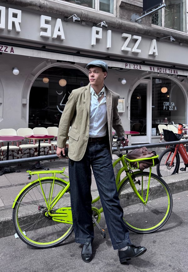 Jeune homme en veste et casquette debout à côté d’un vélo vert vif devant un café avec terrasse extérieure et une enseigne de pizza.