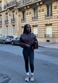 Brun cropped pufferjakke med frontlommer, høy midje svarte leggings, hvite sokker og grå joggesko. Brun håndveske fremhever stilen.