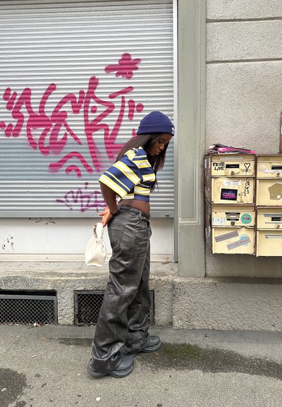 Camisa de rayas cortas en azul y amarillo, pantalones holgados negros y calzado chunky negro. Los accesorios incluyen un bolso blanco y una gorra.