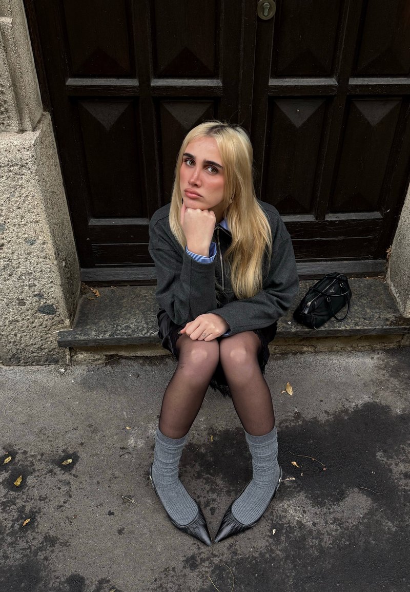 Young woman with long blonde hair, wearing dark jacket, black skirt, gray socks, and black pointed shoes, sitting on stone step by wooden door.