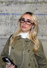 Blond kvinne med briller og ørepropper holder en telefon, kledd i en olivengrønn trenchcoat foran en teksturert betongvegg.