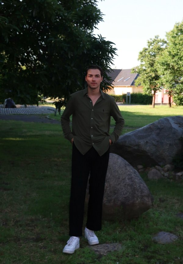 Jeune homme debout sur l'herbe près de grandes roches et d'arbres, portant une chemise foncée, un pantalon noir et des baskets blanches dans un parc ensoleillé.