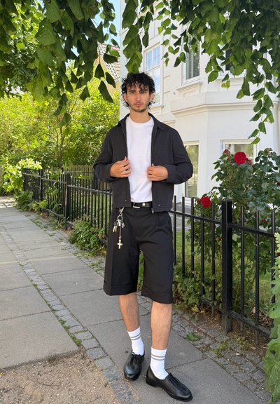 Chaqueta negra con cremallera sobre una camiseta blanca, pantalones cortos negros y zapatos negros. Los accesorios incluyen llaves y una cruz en el llavero.