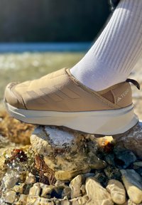 Rudi slip-on bateliai, pagaminti iš tekstūruotos medžiagos, su trimis šoninėmis juostelėmis, storu baltu tarpiniais padu ir rudais guminiais padais.