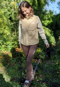 Hellbraune Fleece-Weste mit einer Fronttasche, kombiniert mit dunkelbraunen Leggings mit weißem Logo. Die Person hält eine schwarze Wasserflasche.
