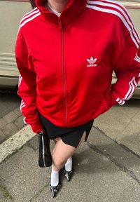 Rdeča Adidas jakna z zadrgo in belimi črtami, črna krila, bele nogavice in črne špicaste čevlje z zaponkami.