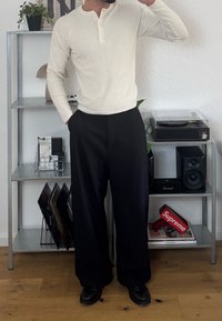 Light beige ribbed long-sleeve shirt with button placket, paired with loose black trousers and black shoes, standing in a contemporary interior.
