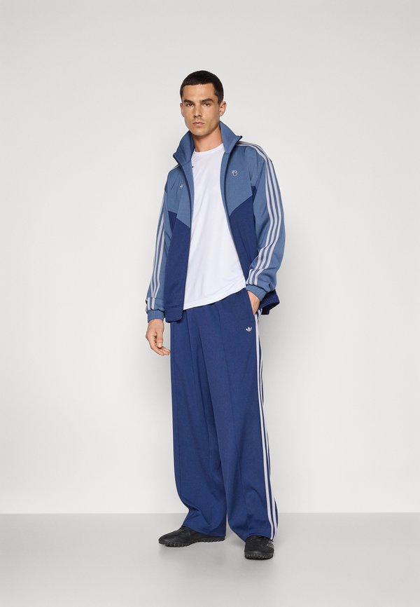 Casaco de treino azul com calças largas a condizer, ambas com três riscas brancas. O casaco apresenta um fecho éclair e um colarinho levantado, combinado com uma camisa branca.