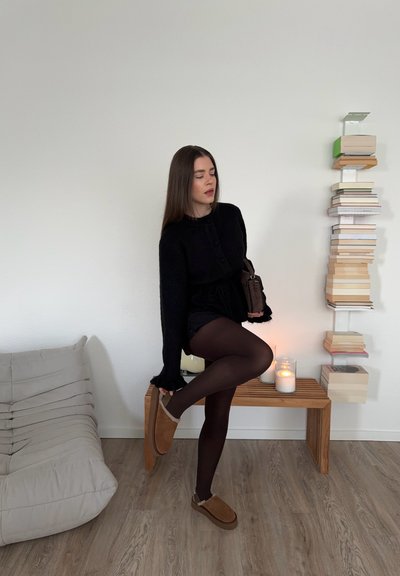 Cárdigan negro de punto con botones, acompañado de pantalones cortos negros y zapatillas de piel de fur con forro marrón. Banco de madera y libros apilados en el fondo.