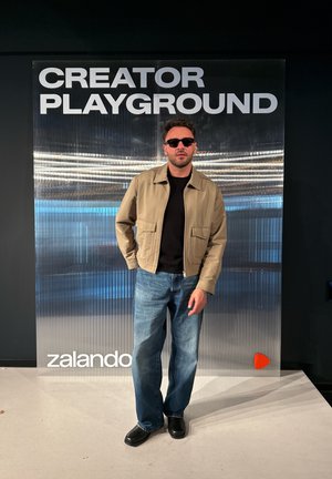 Hombre con gafas de sol, chaqueta beige, camisa negra, vaqueros azules y zapatos negros, de pie frente a un cartel de "CREATOR PLAYGROUND" y Zalando.