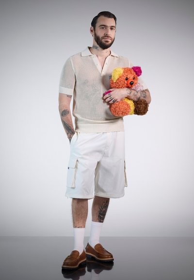 Polo de punto beige, pantalones cortos cargo blancos, mocasines marrones, calcetines blancos. Sosteniendo un oso de peluche colorido con texturas y formas distintas.