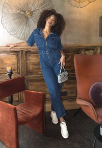 Mono de mezclilla con mangas cortas, cintura ceñida y cremallera frontal; complementado con un pequeño bolso de cuentas. Zapatillas blancas en los pies.