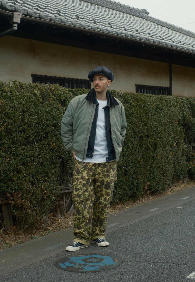 Jeune homme portant une casquette, une veste verte, une chemise blanche et un pantalon camouflage se tient dans une rue à côté de haies taillées et d'une maison traditionnelle au toit en tuiles.