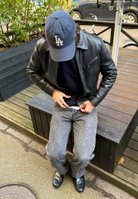 Czarna skórzana kurtka z kieszenią na zamek, granatowa czapka z logo, ciemny sweter, szare dżinsy i czarne mokasyny na drewnianej ławce na zewnątrz.